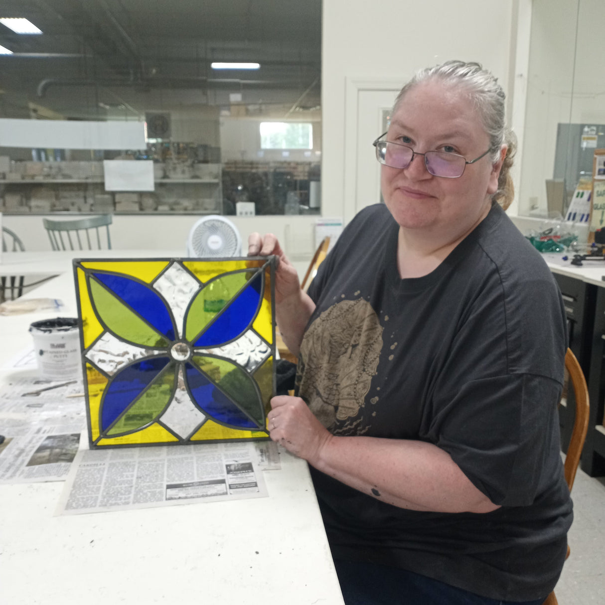 Person holding a stained glass panel with a blue and green design in a workshop setting.