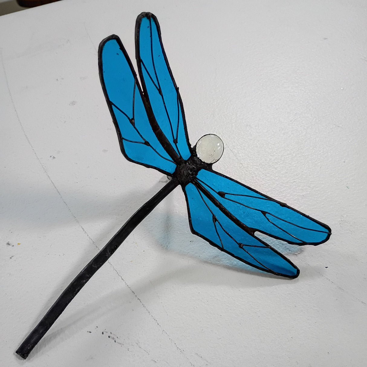 A stained glass dragonfly ornament with bright blue wings, black outlines, and a small white circular detail, resting on a white surface.
