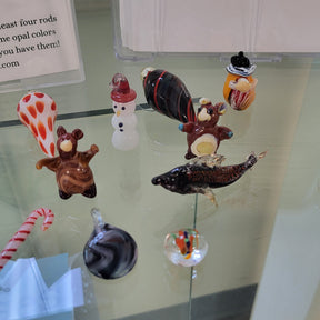 A display of hand-blown glass ornaments and figurines arranged on a glass shelf, including a snowman, a fish, a striped candy cane, and small decorative pieces, with an informational sign in the background.