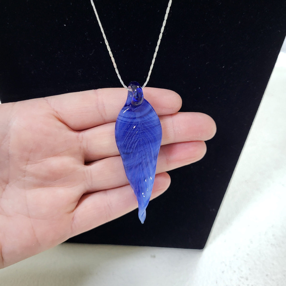 A hand holding a blue teardrop-shaped pendant necklace on a silver chain against a black display background.