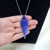 A hand holding a blue teardrop-shaped pendant necklace on a silver chain against a black display background.