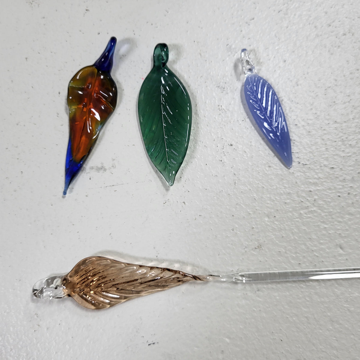 Four hand-blown glass leaf charms in various colors and finishes, including amber, green, blue, and clear glass, arranged on a textured off-white background.