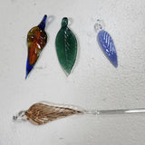 Four hand-blown glass leaf charms in various colors and finishes, including amber, green, blue, and clear glass, arranged on a textured off-white background.