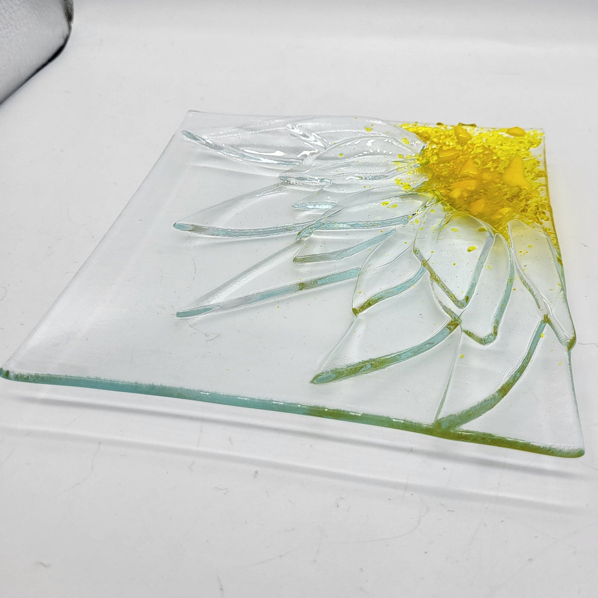 A square clear glass plate featuring an embossed or molded sunflower design with a bright yellow center and pale green to clear petals, photographed on a white studio background.
