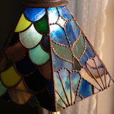 A close-up of a stained glass table lamp shade with a scalloped, fish-scale pattern in shades of blue, green, yellow, brown, and cream, with copper-toned metal leading. The shade is illuminated, casting a warm glow, and is set against a dark background with a white lace curtain on the right.