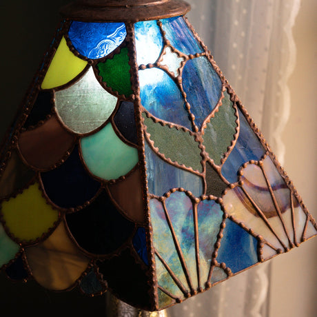 A close-up of a stained glass table lamp shade with a scalloped, fish-scale pattern in shades of blue, green, yellow, brown, and cream, with copper-toned metal leading. The shade is illuminated, casting a warm glow, and is set against a dark background with a white lace curtain on the right.