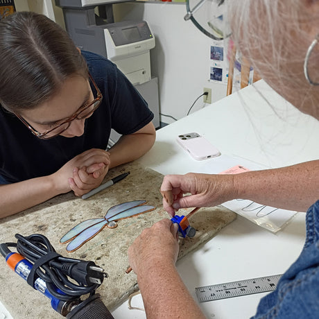 A person in a blue denim shirt uses a small tool on a dragonfly wing frame placed on a workbench, while another person watches closely. The scene is set in a workshop with tools, a phone, and a printer visible in the background.