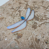 A dragonfly-shaped stained glass piece with iridescent white wings, copper-colored outlines, and a small blue jewel accent, displayed on a textured tan stone surface.