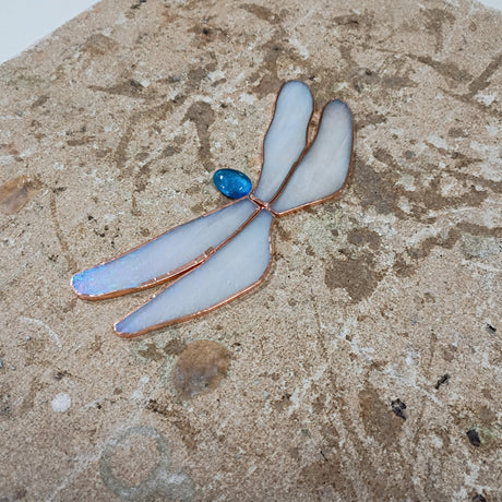 A dragonfly-shaped stained glass piece with iridescent white wings, copper-colored outlines, and a small blue jewel accent, displayed on a textured tan stone surface.