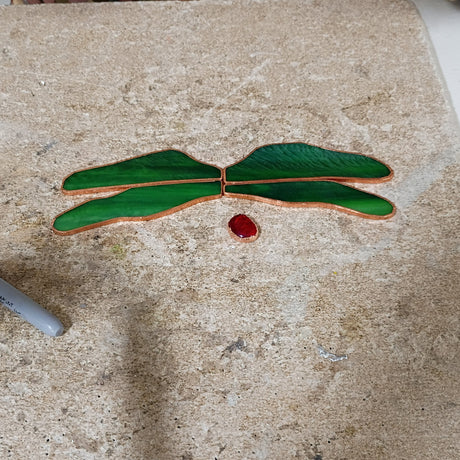 A handmade dragonfly cutout made from green stained glass pieces with copper-colored edges and a small red center, placed on a textured beige workbench with a hot air gun and other tools nearby.